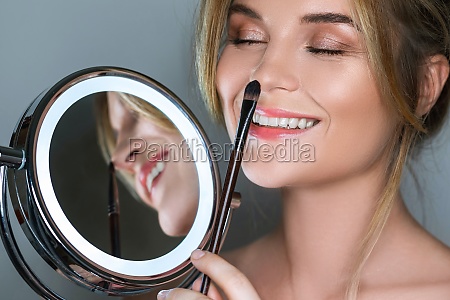 Beautiful woman with make-up brush and round mirror with LED light on gray background